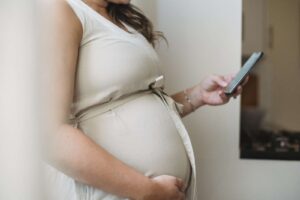 Vue latérale d'une femme enceinte anonyme envoyant des messages texte sur son téléphone, debout dans une pièce, à la lumière du jour.