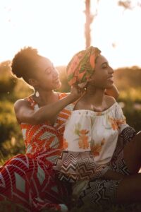 une femme enceinte en robe blanche à fleurs se fait coiffer par une femme. Elles sont assises toutes deux dans l'herbe avec les rayons du soleil