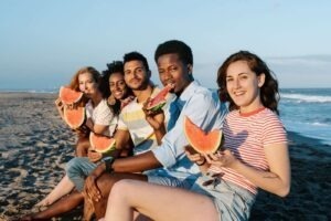 Jeunes amis multiraciaux dégustant des tranches de melon d'eau sur un rivage sablonneux en regardant l'appareil photo.